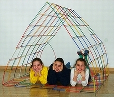 Trzy uśmiechnięte dziewczynki leżące w konstrukcji przypominającej namiot, zbudowanej z kolorowych rurek połączonych ze sobą. Podłoga wyłożona parkietem.